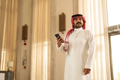 Using a modern technical device, browsing social media on a mobile phone, an Arab Gulf Saudi man wearing a white thobe and a ghutrah stands inside a hotel holding a mobile phone in his hand, looking at something.