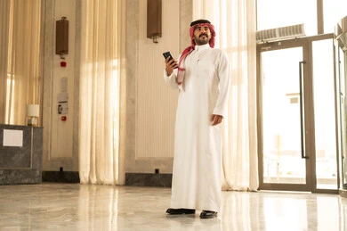 النظر الى شيء ما، تصفح تطبيق الهاتف المحمول، الدردشة مع الاصدقاء عبر الانترنت، رجل عربي خليجي سعودي يرتدي الثوب الابيض و الشماغ يقف داخل الفندق يتابع اخبار وسائل التواصل الاجتماعي باستخدام الجوال