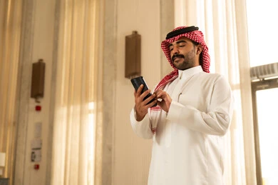 Gestures of admiration and joy, browsing a mobile application, chatting with friends online, an Arab Gulf Saudi man wearing a white thobe and a ghutrah standing inside a hotel following social media news using his mobile phone.