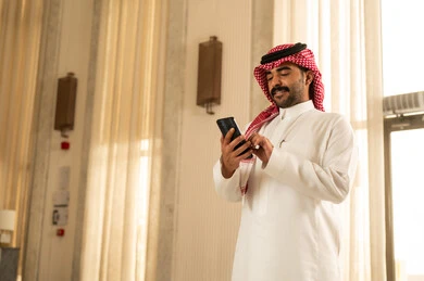 Browsing a mobile application, chatting with friends online, a Saudi Arabian Gulf man wearing a white thobe and a ghutrah stands inside a hotel following social media news using his mobile phone, showing gestures of admiration and joy.