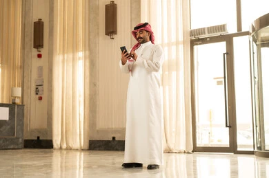 الدردشة مع الاصدقاء عبر الانترنت، رجل عربي خليجي سعودي يرتدي الثوب الابيض و الشماغ يقف داخل الفندق يتابع اخبار وسائل التواصل الاجتماعي باستخدام الجوال، تصفح تطبيق الهاتف المحمول، ايماءات الاعجاب والسرور