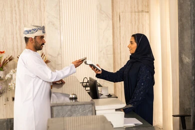 The use of modern means and technologies in electronic payment, an Arab Gulf Omani man wearing a dishdasha and turban in a luxury hotel, the ease of automatic payment in Saudi Arabia, controlling reservations and payments through a smart watch via mobile, enjoying a pleasant time with gestures of happiness and joy.