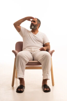 Expressions of trying to see distant things, the concept of looking forward to the future, looking at a place, a portrait of an Arab Saudi Gulf man wearing white pants and a t-shirt sitting on a chair raising his hand to his head with gestures of searching for something specific, white background.