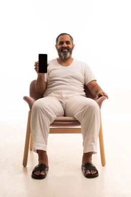 مفهوم التسويق الرقمي الالكتروني، عرض تطبيقات وخدمات الهاتف المحمول،  بورتريه لرجل عربي سعودي خليجي يرتدي سروال وفانيلا أبيض يجلس على كرسي يحمل بيده جوال بشاشة سوداء فارغة، استخدام التكنولوجيا المتطورة، خلفية بيضاء