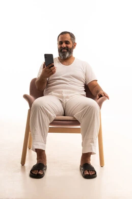 برامج التواصل الاجتماعي عبر الهاتف المحمول، استخدام التطبيقات الحديثة و التسويق الألكتروني, بورتريه لرجل عربي سعودي خليجي يرتدي سروال وفانيلا أبيض يجلس على كرسي يستخدم الجوال, خلفية بيضاء