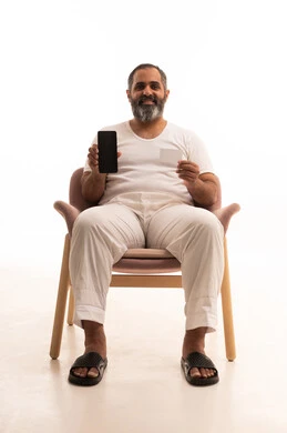 Using mobile applications for payment and shopping, a portrait of an Arab Saudi Gulf man wearing white pants and a t-shirt sitting on a chair holding an electronic card and a mobile phone, looking with gestures of cheerfulness and joy, the possibility of electronic payment through advanced modern technologies, white background.