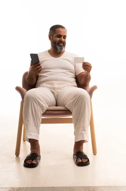 الدفع الإلكتروني عبر التقنيات الحديثة المتطورة، استخدام تطبيقات الجوال للدفع والشراء، بورتريه لرجل عربي سعودي خليجي يرتدي سروال وفانيلا أبيض يجلس على كرسي يمسك بيده البطاقة الإلكترونية والهاتف المحمول، خلفية بيضاء