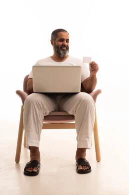 Online electronic payment, looking at the camera with gestures of joy using modern means, a portrait of an Arab Saudi Gulf man wearing white pants and a t-shirt sitting on a chair holding a laptop and a credit card in his hand, white background.