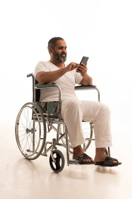 Communicating with family and friends, making a video call via mobile, browsing social media, using modern technology, and a portrait of an Arab Saudi Gulf man wearing white pants and a t-shirt sitting in a wheelchair working on a mobile phone, with a white background.
