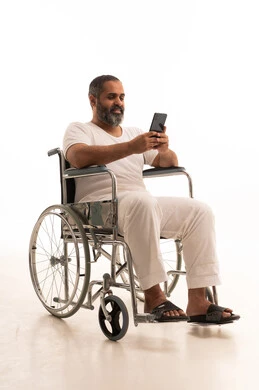 Making a video call via mobile, communicating with family and friends, browsing social media, using modern technology, and a portrait of an Arab Saudi Gulf man wearing white pants and a t-shirt sitting in a wheelchair working on a mobile phone, with a white background.