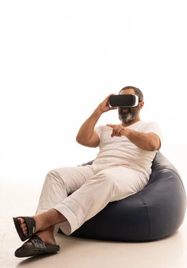 Attempting to click on something, a portrait of an Arab Saudi Gulf man wearing white pants and a t-shirt sitting on a chair between bags, wearing 3D glasses, raising his index finger upwards, pointing to a place with expressions of immersion and simulating the imaginary world, white background.
