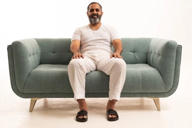 النظر الى الكاميرا  بايماءات السعادة والسرور، الاستمتاع و الاسترخاء بالأجواء الهادئة، بورتريه لرجل عربي سعودي خليجي يرتدي سروال وفانيلا أبيض يجلس على الاريكة، خلفية بيضاء