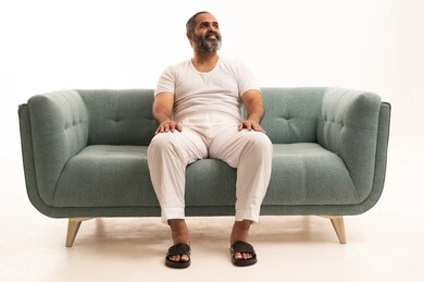 الاستمتاع و الاسترخاء بالأجواء الهادئة، بورتريه لرجل عربي سعودي خليجي يرتدي سروال وفانيلا أبيض يجلس على الاريكة ينظر الى شيء ما بايماءات السعادة والسرور، خلفية بيضاء