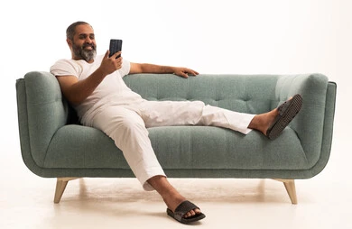 استخدام جهاز تقني حديث، الدردشة مع الأهل والأصدقاء،  بورتريه لرجل عربي سعودي خليجي يرتدي سروال وفانيلا أبيض يجلس على الاريكة يمسك بيده الهاتف المحمول، دمج التقنية والتكنولوجيا بالحياة اليومية، تصفح مواقع التواصل الاجتماعي، خلفية بيضاء