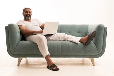 Completing remote work tasks, using modern technologies in administrative work, making video calls, a portrait of an Arab Saudi Gulf man wearing white pants and a t-shirt sitting on the couch and working on a laptop, integrating advanced technology into daily life, white background.