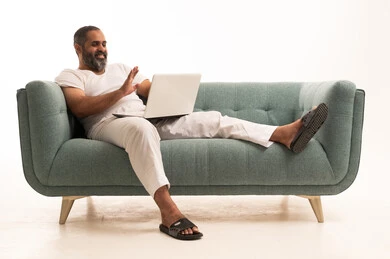 Raising the hand with expressions of peace and greeting, making a video call, working remotely, using advanced modern technology in professional life, a portrait of an Arab Saudi Gulf man wearing white pants and a t-shirt sitting on the sofa working on a laptop, white background.