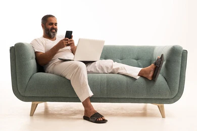 The use of modern and advanced technological devices to complete projects, browsing social media, and working remotely, a portrait of an Arab Saudi Gulf man wearing pants and a white t-shirt sitting on the couch using a phone and a laptop.
