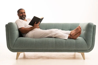 Looking at the camera with expressions of joy, utilizing time for education, the concept of literacy and learning, future planning for goals and ambitions, a portrait of an Arab Saudi Gulf man wearing white pants and a t-shirt sitting on a sofa holding a book that he is reading attentively, with a white background.