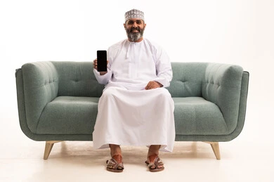 عرض شاشة سوداء فارغه عبر الجوال، بورتريه لرجل عربي خليجي عماني يرتدي الدشداشة و الكمة يمسك بيده الهاتف المحمول، دمج التقنية والتكنولوجيا بالحياة اليومية النظر للكاميرا بتعابير السرور، خلفية بيضاء