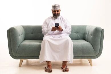 Using a modern technical device, chatting with family and friends, a portrait of an Arab Gulf Omani man wearing a dishdasha and kumma holding a mobile phone, integrating technology into daily life, browsing social media, white background.
