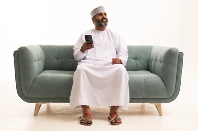 الدردشة مع الأهل والأصدقاء، استخدام جهاز تقني حديث، بورتريه لرجل عربي خليجي عماني يرتدي الدشداشة و الكمة يمسك بيده الهاتف المحمول، دمج التقنية والتكنولوجيا بالحياة اليومية، تصفح مواقع التواصل الاجتماعي، خلفية بيضاء