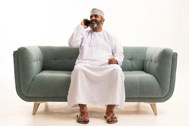 الرد على الاتصالات والمكالمات، التواصل مع الأصدقاء والاقارب، استخدام التقنيات الحديثة المتطورة، سماع أخبار سعيدة عبر الهاتف المحمول، بورتريه لرجل عربي خليجي عماني يرتدي الدشداشة و الكمة يجلس على اريكه يقوم بإجراء مكالمة هاتفية بايماءات السعاده، خلفيه بيضاء