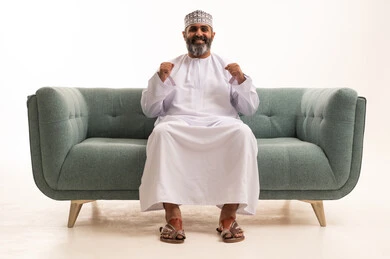 مفهوم الأندية والتشجيع، متابعة المباراة و دعم وتشجيع المنتخب , بورتريه لرجل عربي خليجي عماني يرتدي الدشداشة و الكمة يجلس على اريكه يشاهد مباراة للدوري العربي في المنزل, ايماءات وجه تدل على النصر، خلفية بيضاء