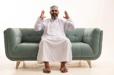 متابعة المباراة و دعم وتشجيع المنتخب  , بورتريه لرجل عربي خليجي عماني يرتدي الدشداشة و الكمة يجلس على اريكه يشاهد مباراة للدوري العربي في المنزل, ايماءات وجه تدل على النصر , مفهوم الأندية والتشجيع، خلفية بيضاء