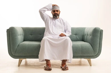 أنديه رياضية مختلفة، بورتريه لرجل عربي خليجي عماني يرتدي الدشداشة و الكمة يجلس على اريكه يضع يده على رأسه بتعابير الحزن والتفاعل مع المنتخب، إيماءات الخسارة والفشل، مشاهدة ومتابعة مباراة كرة القدم،خلفية بيضاء