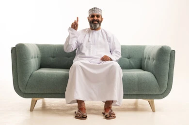 الاستفسار والاشارة إلى شيء ما, بورتريه لرجل عربي خليجي عماني يرتدي الدشداشة و الكمة يجلس على اريكه يرفع اصبعه للأعلى بإيماءات التوضيح، النظرإلى الكاميرا بتعابير الاستفهام والتساؤل، خلفية بيضاء
