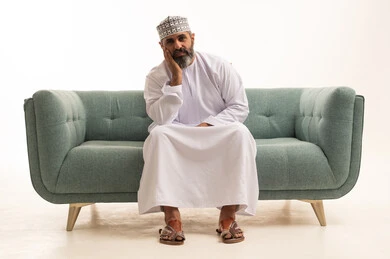 The concept of oppression and loss, regret and sorrow over something, a portrait of an Arab Gulf Omani man wearing a dishdasha and kumma sitting on a sofa with his hand on his chin in gestures of sadness, feeling of discomfort and disappointment, white background.