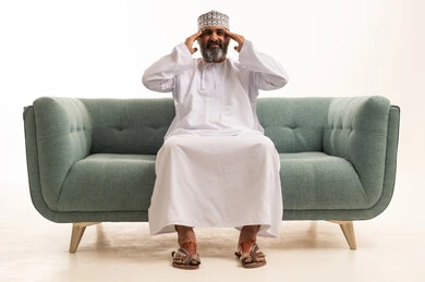 الحاجه للعلاج وطلب المساعده, بورتريه لرجل عربي خليجي عماني يرتدي الدشداشة و الكمة يجلس على اريكه يمسك رأسه بيديه من شدة الألم, الشعور بالتعب والإرهاق, الإصابة بنوبات الشقيقة، إيماءات الصداع والإحساس بالمرض, خلفية بيضاء
