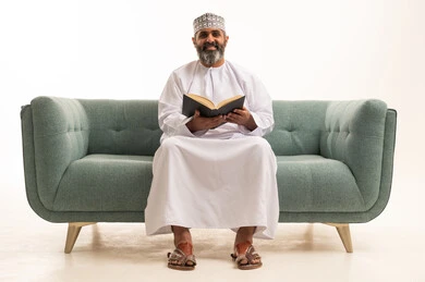 The concept of literacy, a hobby of love for reading and exploration, spending free time learning and cultural activities, a portrait of an Arab Gulf Omani man wearing a dishdasha and a kumma sitting on a sofa holding a scientific educational book, white background.