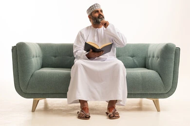Gestures of thinking about something, a hobby of love for reading and exploration, spending free time learning and culture, the concept of literacy, a portrait of an Arab Gulf Omani man wearing a dishdasha and kumma sitting on a sofa holding a scientific educational book in his hand, white background.