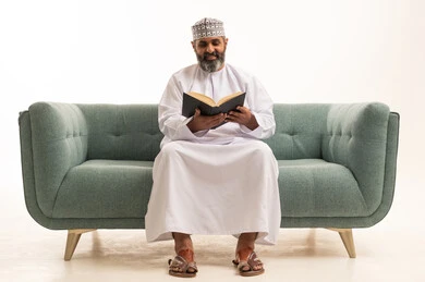 هواية حب المطالعة و الاستكشاف، القراءة بتركيز وتمعن, قضاء وقت الفراغ بالتعلم والثقافة، مفهوم محو الامية، بورتريه لرجل عربي خليجي عماني يرتدي الدشداشة و الكمة يجلس على اريكه يحمل بيده كتاب علمي تثقيفي، خلفية بيضاء