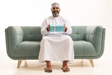 Expressing love, appreciation, and respect, giving gifts on holidays and special occasions, the concept of gratitude and thankfulness, a portrait of an Arab Gulf Omani man wearing a dishdasha and kumma sitting on a sofa holding a gift box in his hands, white background.