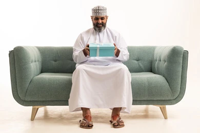 Gift-giving during holidays and special occasions, the concept of gratitude and appreciation, expressing love, respect, and admiration, a portrait of an Arab Gulf Omani man wearing a dishdasha and kumma sitting on a sofa holding a gift box in his hands, white background.