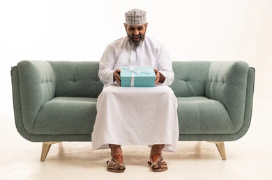 مفهوم الشكر والامتنان، تقديم الهدايا بالاعياد والمناسبات الخاصة، التعبير عن الحب والتقدير والاحترام،  بورتريه لرجل عربي خليجي عماني يرتدي الدشداشة و الكمة يجلس على اريكه يحمل بيديه صندوق هدية , خلفية بيضاء