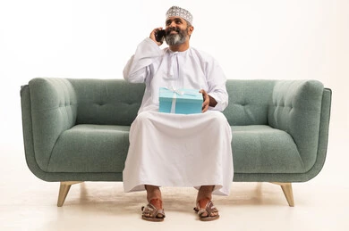 التواصل مع الأصدقاء والاقارب، سماع أخبار سارَة عبر الهاتف المحمول، بورتريه لرجل عربي خليجي عماني يرتدي الدشداشة و الكمة يجلس على اريكه يقوم بإجراء مكالمة هاتفية ويحمل بيده علبة هدية بإيماءات السرور، أعياد ومفاجآت ومناسبات سعيدة، خلفية بيضاء