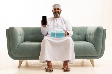 عرض شاشة سوداء فارغه عبر الهاتف المحمول، بورتريه لرجل عربي خليجي عماني يرتدي الدشداشة و الكمة يجلس على اريكه يمسك بيدية الجوال و علبة هدية بإيماءات السرور، أعياد ومفاجآت ومناسبات سعيدة، خلفية بيضاء