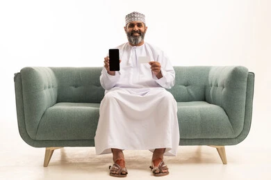 عرض شاشة سوداء فارغه عبر الجوال، استخدام تطبيقات الموبيل للدفع والشراء، بورتريه لرجل عربي خليجي عماني يرتدي الدشداشة و الكمة يجلس على اريكه يمسك بيده البطاقة الإلكترونية والهاتف المحمول ينظر بإيماءات البشاشة والسرور، إمكانية الدفع الإلكتروني عبر التقنيات الحديثة المتطورة، خلفية بيضاء