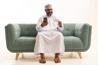 الدفع والشراء من خلال برامج وتطبيقات الجوال، التقنيات الحديثة والمتطورة، بورتريه لرجل عربي خليجي عماني يرتدي الدشداشة و الكمة يجلس على اريكه يمسك بيده الهاتف المحمول ينظر الى بطاقة الإئتمان البنكية بإيماءات الفرح و السرور، خلفية بيضاء