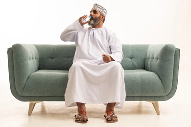 المحافظة على سلامة الجهاز المناعي، التخلص من سموم الجسم، أهمية شرب الماء النقي للمحافظة على بشرة نضرة، بورتريه لرجل عربي خليجي عماني يرتدي الدشداشة و الكمة يجلس على اريكه يقوم بشرب المياه من عبوة بلاستيكية، الحفاظ على صحة الكلى ومعالجتها، خلفية بيضاء