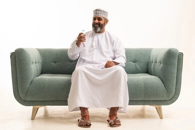 التخلص من سموم الجسم، أهمية شرب الماء النقي للمحافظة على بشرة نضرة، بورتريه لرجل عربي خليجي عماني يرتدي الدشداشة و الكمة يجلس على اريكه يقوم بشرب المياه من عبوة بلاستيكية، المحافظة على سلامة الجهاز المناعي، الحفاظ على صحة الكلى ومعالجتها، خلفية بيضاء