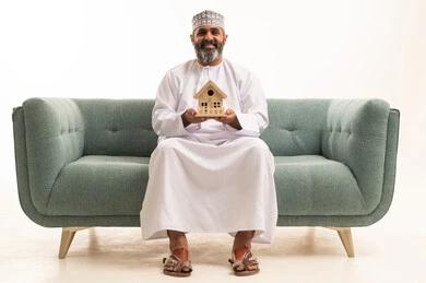 بيع وشراء العقارات، إيماءات السرور والسعادة، السعي لامتلاك بيت جديد، بورتريه لرجل عربي خليجي عماني يرتدي الدشداشة و الكمة يجلس على اريكه يحمل بيديه مجسم خشبي صغير لمنزل ، خلفية بيضاء