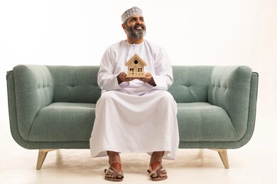 النظر الى شيء ما،  بيع وشراء العقارات، إيماءات السرور والسعادة، السعي لامتلاك بيت جديد، بورتريه لرجل عربي خليجي عماني يرتدي الدشداشة و الكمة يجلس على اريكه يحمل بيديه مجسم خشبي صغير لمنزل ، خلفية بيضاء