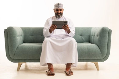 تصفح مواقع التواصل الاجتماعي عبر الجهاز اللوحي، استعمال التكنولوجيا والتقنية الحديثة، المحادثة مع الأهل والأصدقاء، بورتريه لرجل عربي خليجي عماني يرتدي الدشداشة و الكمة يجلس على اريكه يحمل بيده جهاز التابلت، خلفية بيضاء