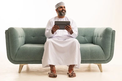 النظر الى شيء ما، استعمال التكنولوجيا والتقنية الحديثة, تصفح مواقع التواصل الاجتماعي عبر الجهاز اللوحي، المحادثة مع الأهل والأصدقاء، بورتريه لرجل عربي خليجي عماني يرتدي الدشداشة و الكمة يجلس على اريكه يحمل بيده جهاز التابلت، خلفية بيضاء