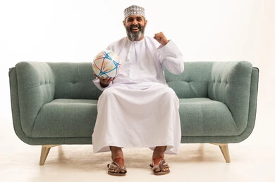 ايماءات السعادة بالانتصار والفوز، أندية وتشجييع منتخبات, بورتريه لرجل عربي خليجي عماني يرتدي الدشداشة و الكمة يجلس على اريكه يقوم بالتفاعل والاندماج مع الفريق يمسك بيدية كره قدم , الشغف في متابعه الفرق، خلفية بيضاء