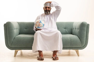 إيماءات الخسارة والفشل، مشاهدة ومتابعة مباراة كرة القدم،بورتريه لرجل عربي خليجي عماني يرتدي الدشداشة و الكمة يجلس على الاريكة يمسك كره القدم ويضع يده على رأسه بتعابير الحزن والتفاعل مع المنتخب، خلفية بيضاء